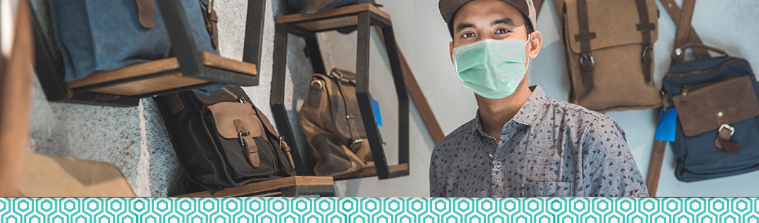 A person wearing a face mask stands inside a retail store displaying backpacks and bags on wall-mounted shelves. The image highlights a small business shopping environment with a variety of handcrafted bags and accessories on display.