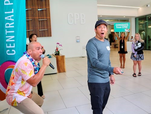 A person stands in the Central Pacific Bank lobby reacting during a surprise celebration while another person speaks into a microphone nearby. A screen in the background reads “Congrats from CPB!” as several attendees look on in the bright, modern space.