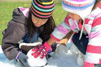 Two children wearing colorful winter hats and gloves work together to build a snowman outdoors.