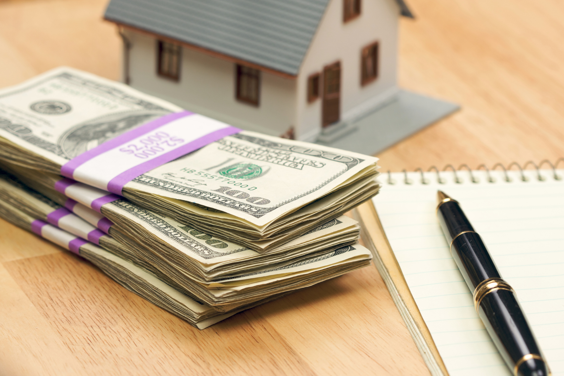 Bundles of U.S. currency, a model house, and a notebook with a pen illustrating home financing, mortgage planning, or real estate investment.