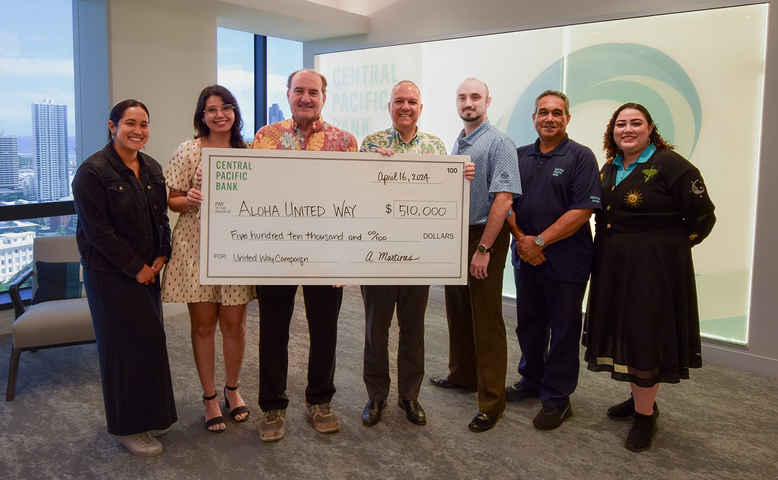 A group of eight people stands indoors holding a large $510,000 Central Pacific Bank donation check presented to Aloha United Way. The check presentation takes place in a conference room with city views and Central Pacific Bank branding in the background.