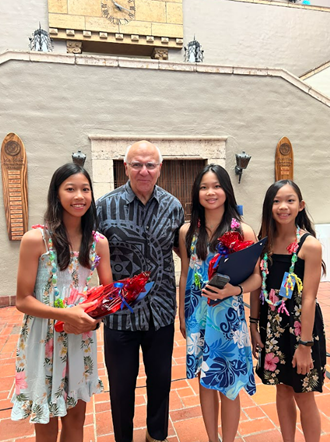 A person stands with three children in an open-air courtyard with tiled flooring and decorative wall plaques. Two of the children hold wrapped gifts and one holds a plaque, posing together in front of a stucco wall with architectural details in the background.