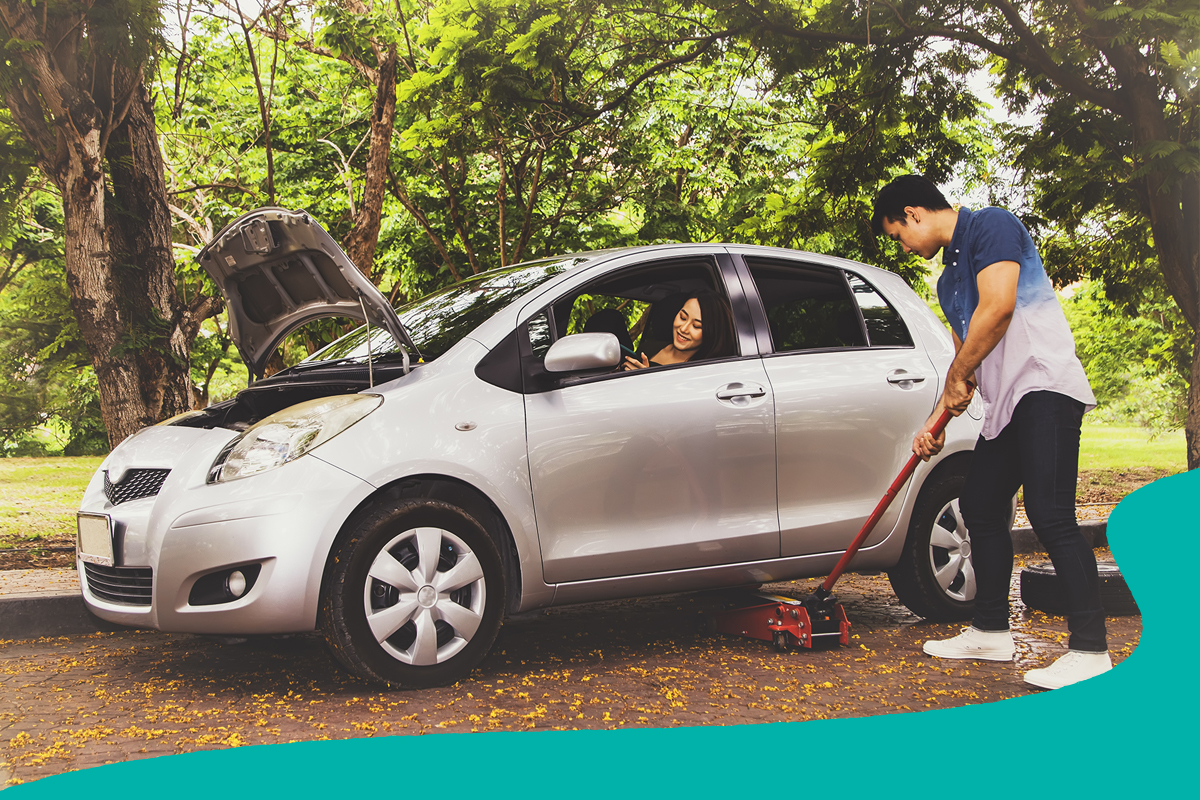 Person using a floor jack to lift a car with the hood open while another person sits in the driver's seat, parked beneath leafy trees.