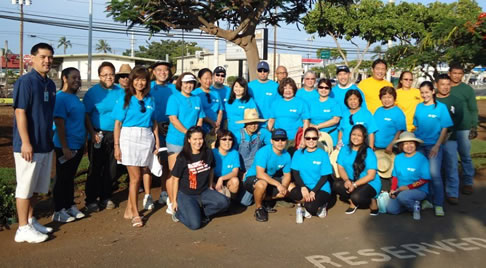 A large group of CPB volunteers wearing matching blue shirts pose for a group photo outdoors in a park-like setting, with trees and a roadway visible in the background.