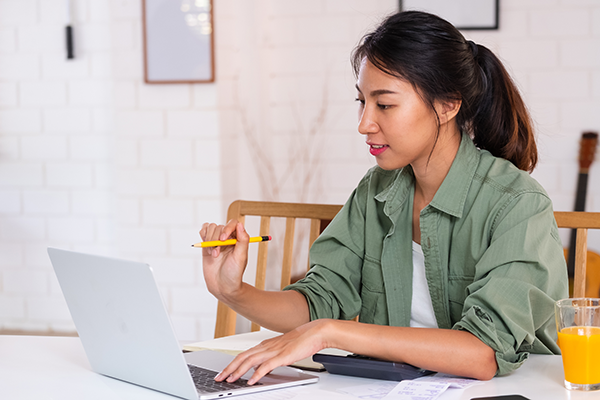 Person reviewing finances at a home office while using a laptop, calculator, and paperwork, with a glass of orange juice on the table.