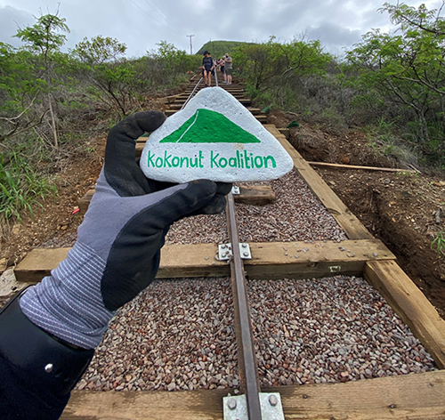 A gloved hand holds a painted rock displaying the Kokonut Koalition logo in the foreground, with a newly constructed hiking trail and stairway ascending a hillside in the background. Several volunteers stand farther up the trail surrounded by native vegetation.