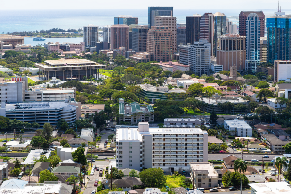 Aerial view of downtown Honolulu with high-rise buildings, residential neighborhoods, green parks, and the Pacific Ocean in the background.