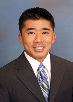 A professional headshot of a person wearing a dark business suit and striped tie, photographed against a blue-gray studio background.