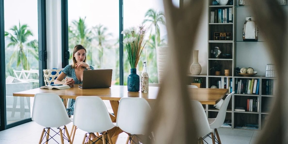 Person working on a laptop at a dining table in a bright home with large windows overlooking palm trees.
