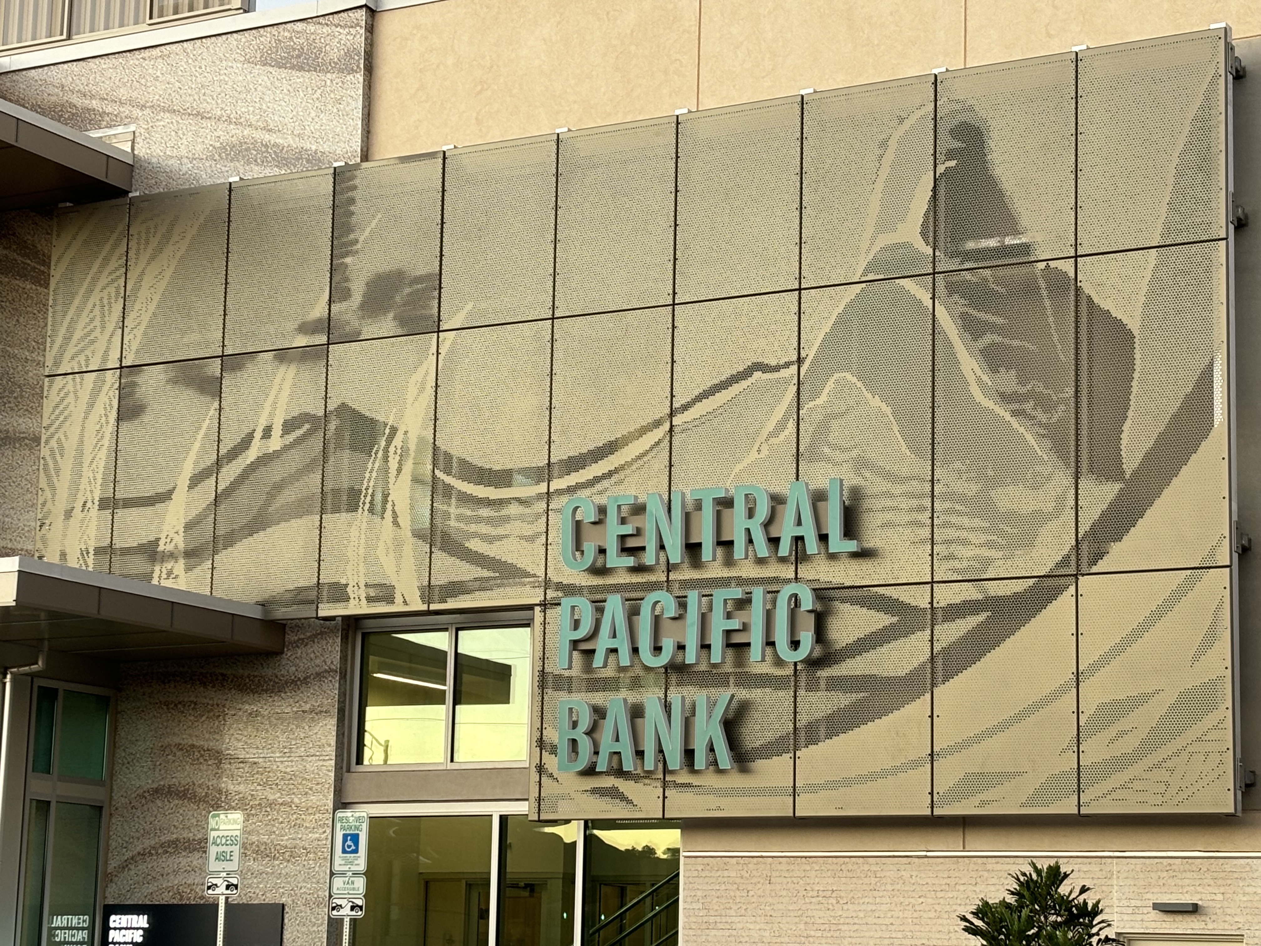 A Central Pacific Bank building façade features large teal lettering and a decorative metal panel displaying an abstract design. The bank entrance, windows, and contemporary architectural details are visible beneath the prominent signage.