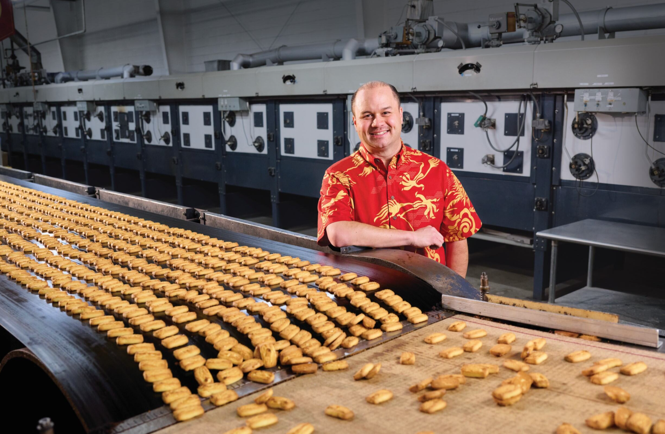 Person in a red patterned aloha shirt stands beside a conveyor belt filled with freshly baked cookies in a commercial bakery or food manufacturing facility.