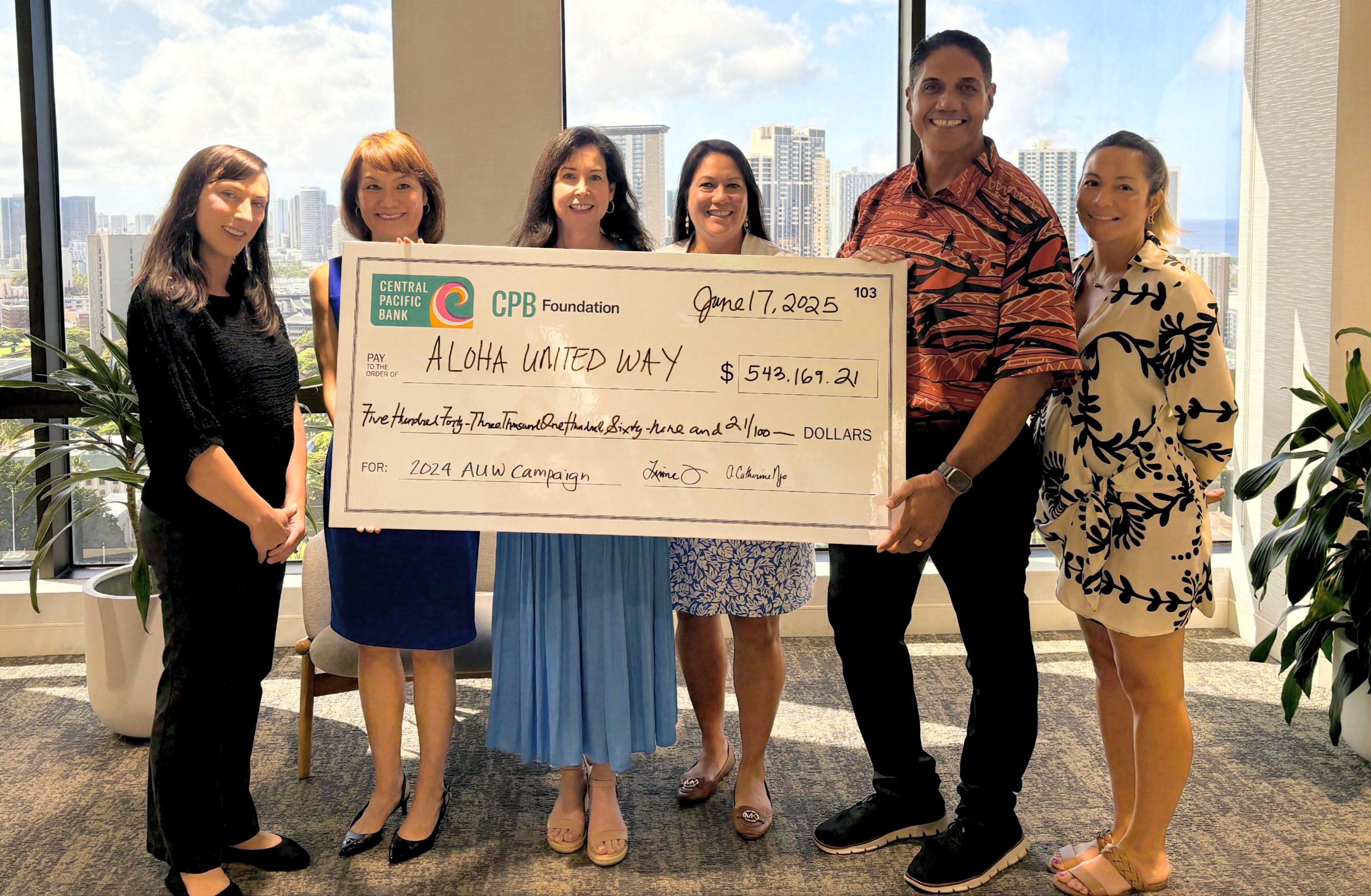 Six people stand in an office with city skyline views behind them, holding a ceremonial check from the CPB Foundation made payable to Aloha United Way for $543,169.21. The check is dated June 17, 2025, and notes the donation is for the 2024 AUW Campaign.