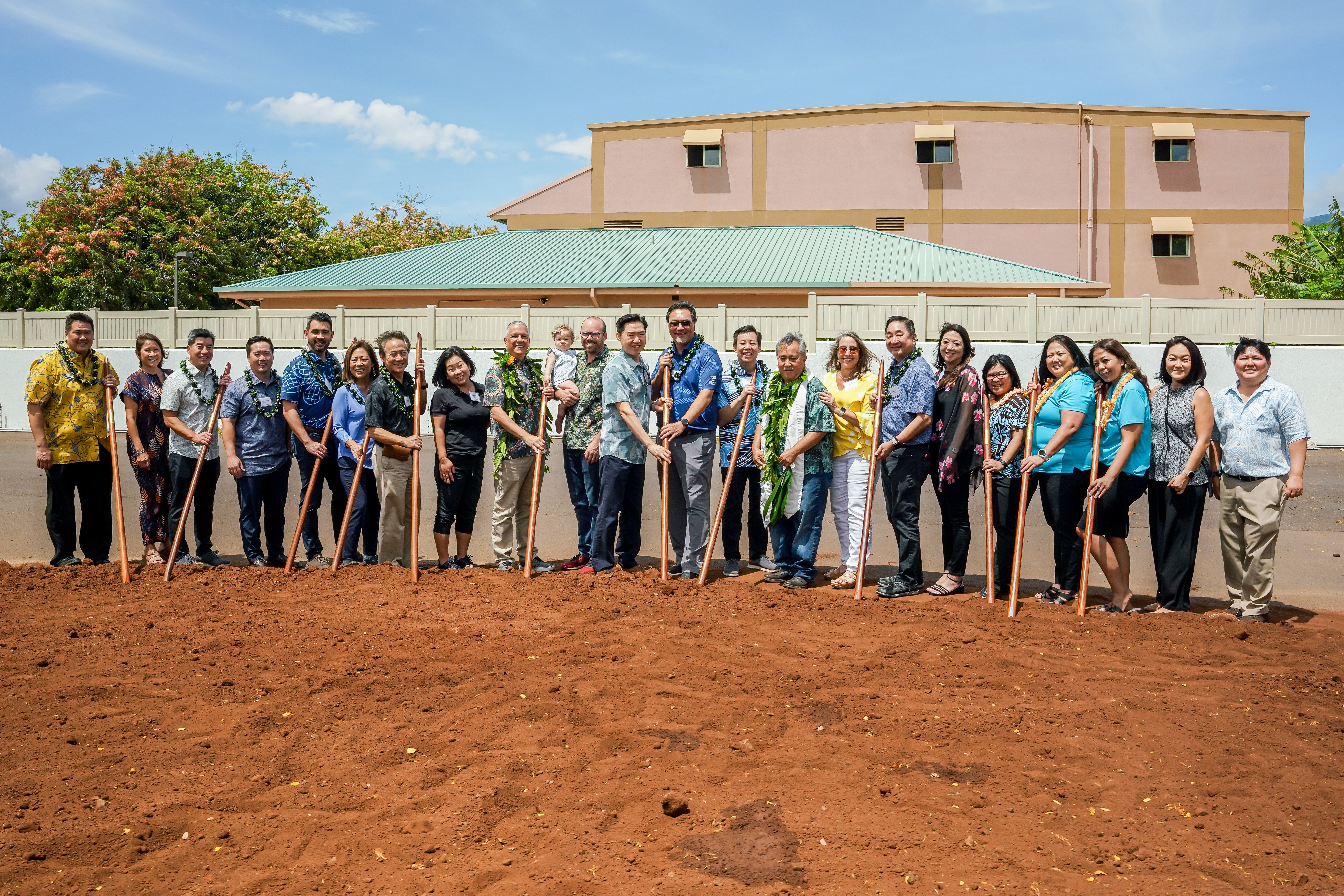 A group of community and business leaders wearing leis hold ceremonial shovels during a groundbreaking event on a cleared construction site. The participants stand in a line on red soil in front of nearby buildings, marking the start of a new development project.
