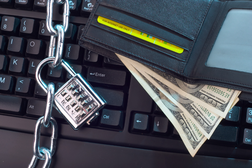 Close-up of a wallet containing cash and a payment card resting on a keyboard next to a chain and combination padlock, illustrating the protection of financial information and online transactions.