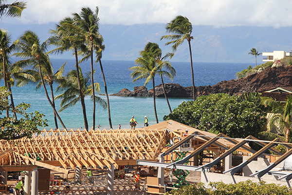 A coastal construction site with partially framed buildings overlooks a tropical shoreline lined with palm trees and blue ocean waters. Workers and construction equipment are visible among the structures, with a scenic island landscape stretching across the background.