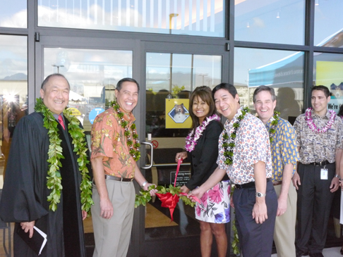 A group of people wearing leis participate in a ribbon-cutting ceremony outside a building entrance, standing near glass doors decorated with a large red bow.