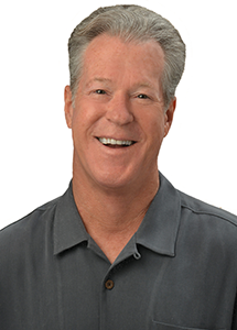 A professional headshot of a person with gray hair wearing a dark gray collared shirt, photographed against a light gray background.