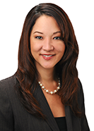 A professional headshot of a person wearing a dark blazer over a black top and a pearl necklace, photographed against a light gray background.