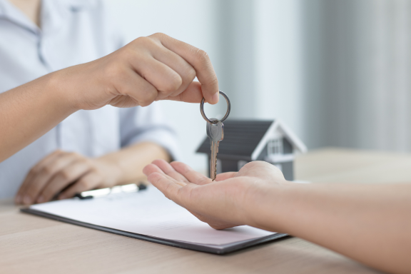 Close-up of a person handing over a house key to another person, with a model home and documents on the desk, representing a home purchase or mortgage closing.