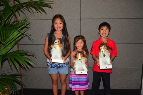 Three children stand indoors holding dog-shaped trophies or awards, posing in front of a gray wall with a potted plant visible to the side.