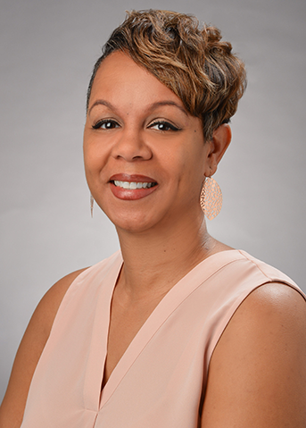 A professional headshot of a person wearing a light pink sleeveless blouse and decorative earrings, photographed against a gray studio background.