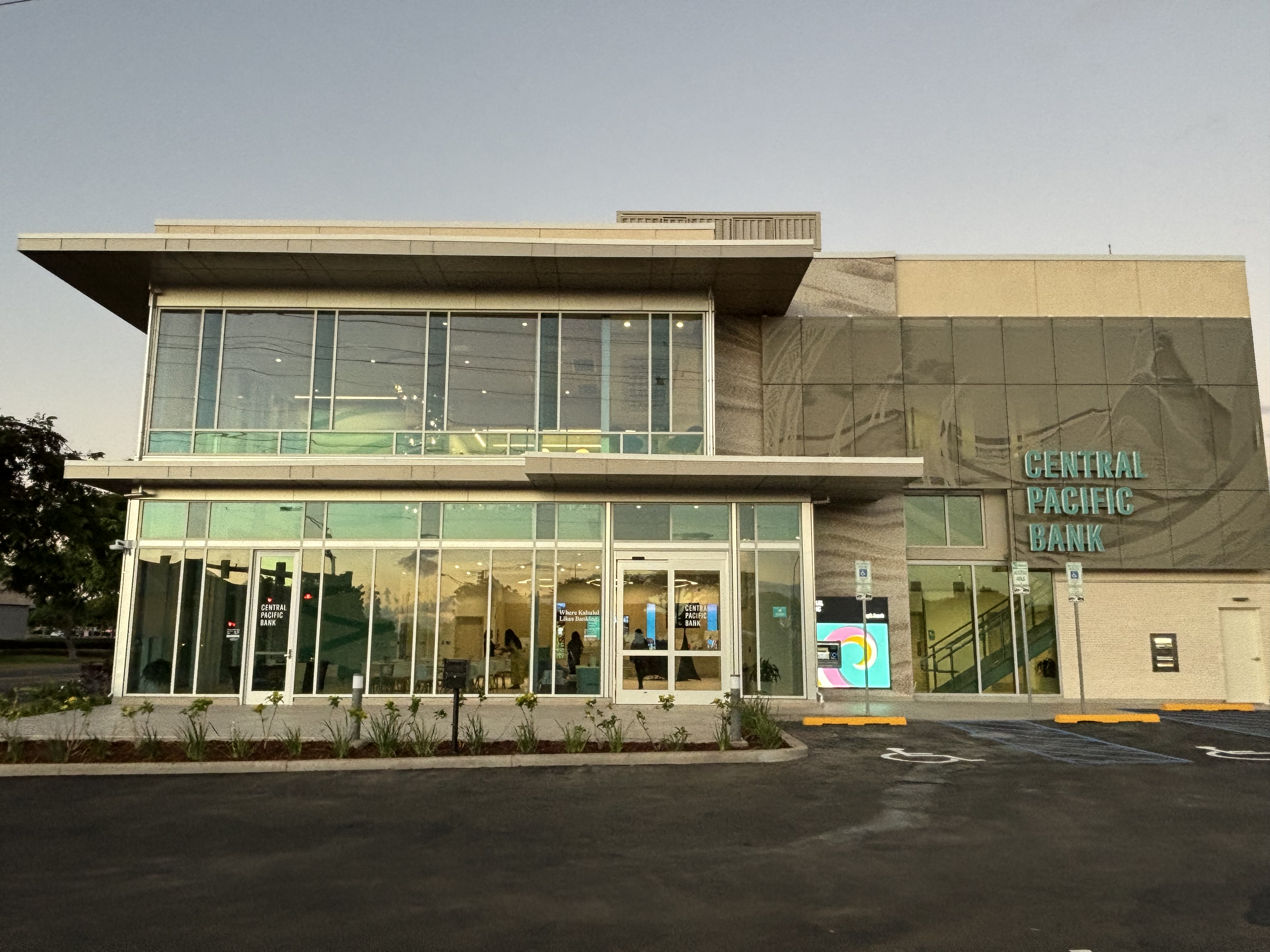 A modern two-story Central Pacific Bank branch with large glass windows, contemporary architecture, and prominent teal signage. The building overlooks a newly paved parking area, with the brightly lit interior visible through the glass façade.