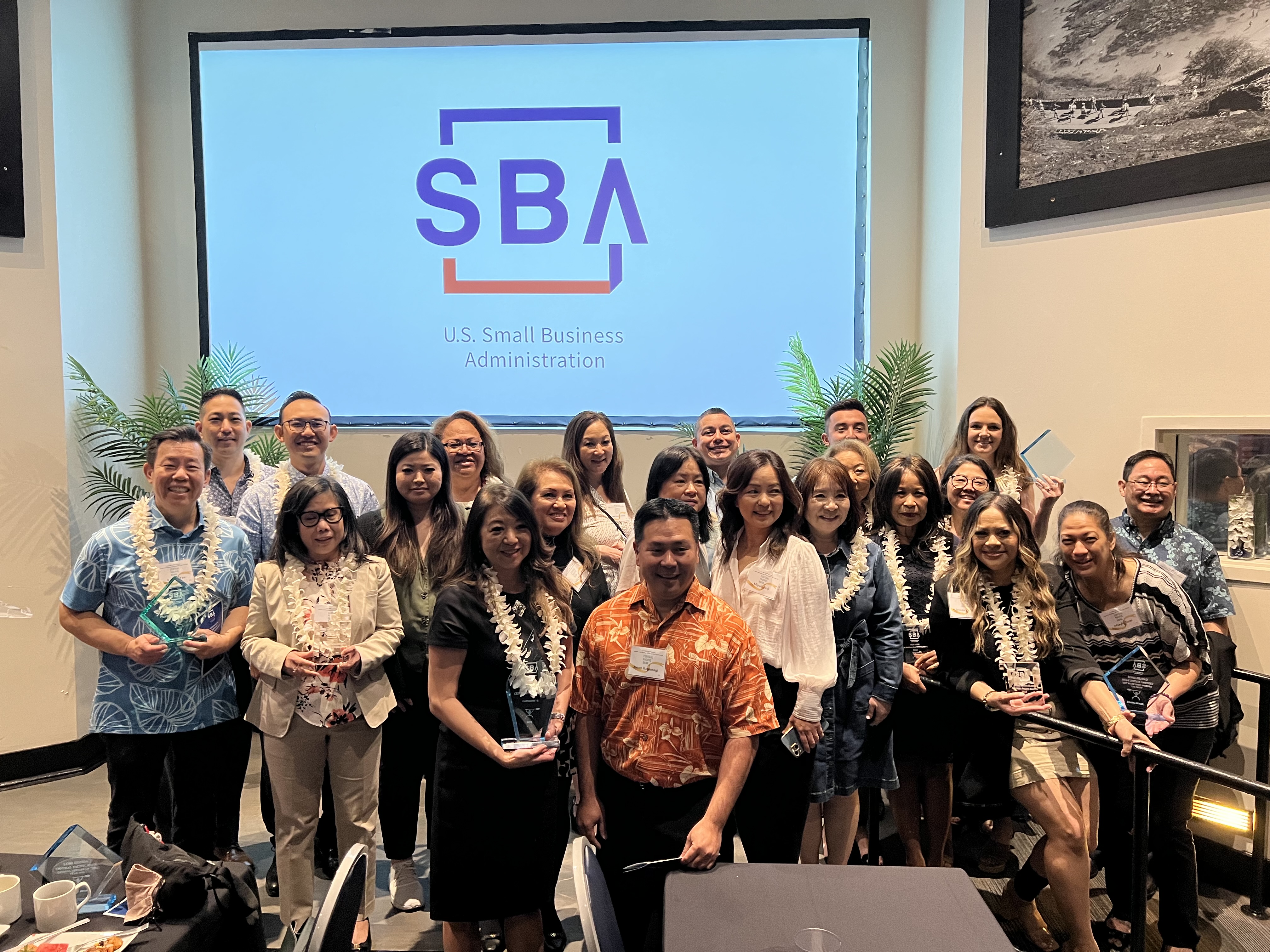 A group of professionals wearing leis pose for a photo at an SBA event, standing in front of a large screen displaying the U.S. Small Business Administration logo. Several participants hold award plaques, highlighting a recognition or awards ceremony.