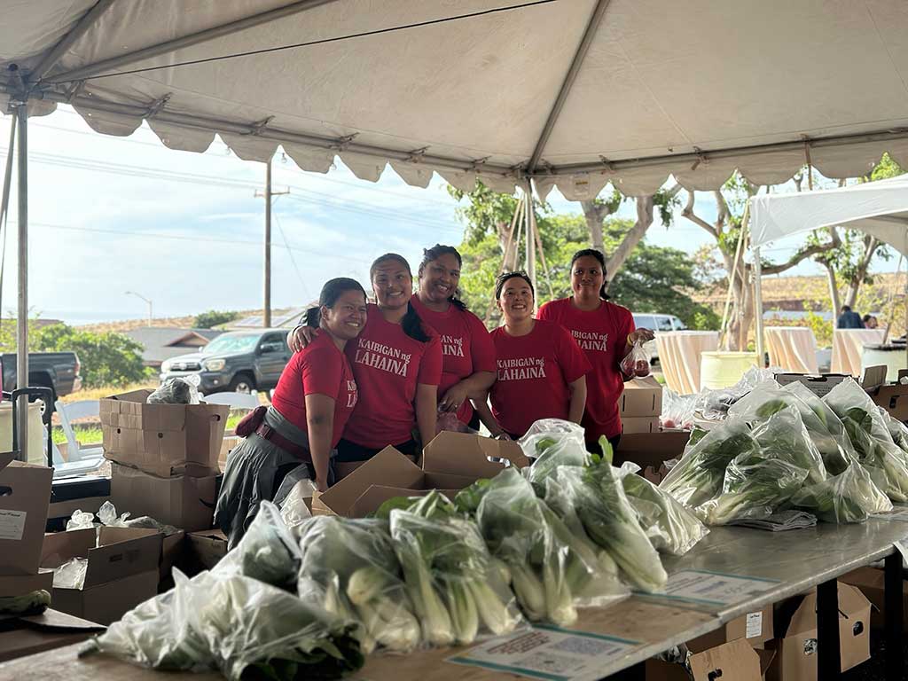A group of volunteers wearing matching red “Kaibigan ng Lahaina” shirts pose behind tables filled with bagged fresh produce under a large tent. The volunteers help organize and distribute food supplies at a community support event.