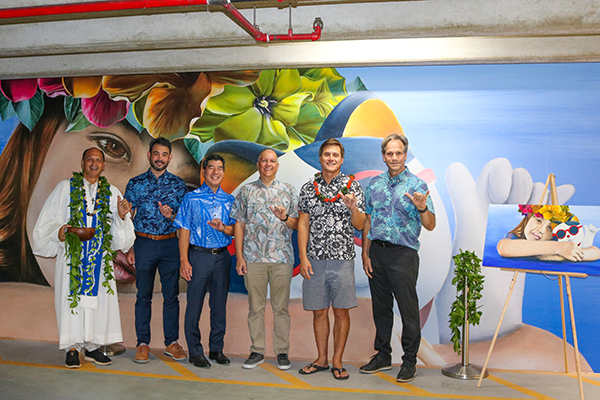A group of six people wearing aloha attire and leis pose in front of a colorful tropical mural featuring large flowers, ocean-themed elements, and a beach scene. The gathering takes place in an indoor parking or event space, with everyone making shaka gestures beside a framed artwork display.