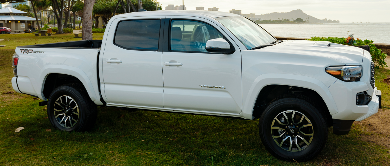 A white Toyota Tacoma pickup truck is parked on a grassy area beside the shoreline, with Diamond Head and the Honolulu skyline visible in the distance. The truck is shown in profile, highlighting its four-door cab and TRD Sport trim.
