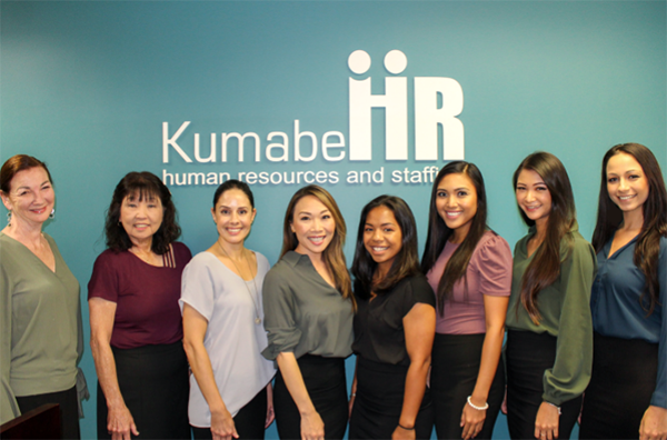 A group of eight employees pose in front of a wall displaying the “Kumabe HR” logo and the words “human resources and staffing.” The team stands together in business attire inside an office, highlighting a professional staffing and human resources organization.