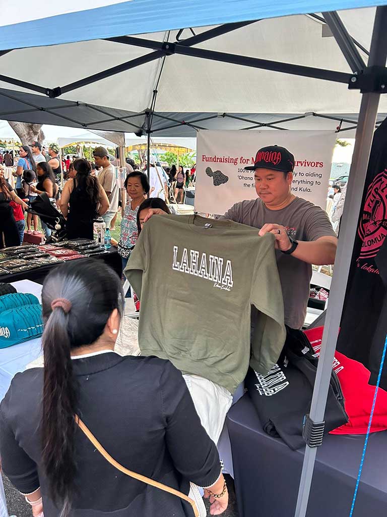 A person displays a sweatshirt reading “LAHAINA” at a community fundraising booth supporting Maui wildfire survivors. Clothing and merchandise are showcased under a canopy tent while visitors browse the event in the background.
