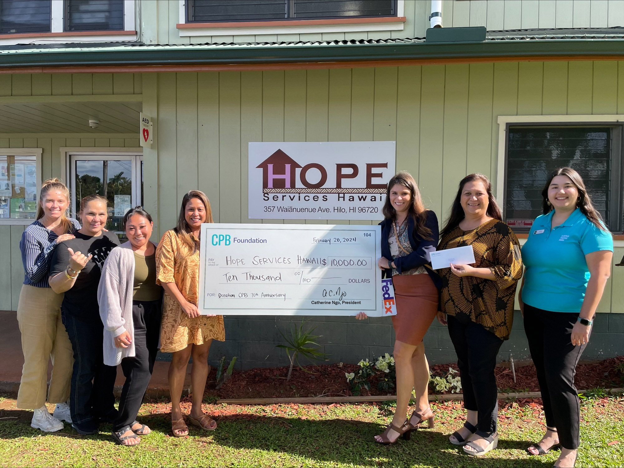 A group of seven people stands outside the HOPE Services Hawaiʻi facility holding a $10,000 CPB Foundation donation check. The presentation takes place in front of the organization’s sign in Hilo, recognizing support for community services and programs.