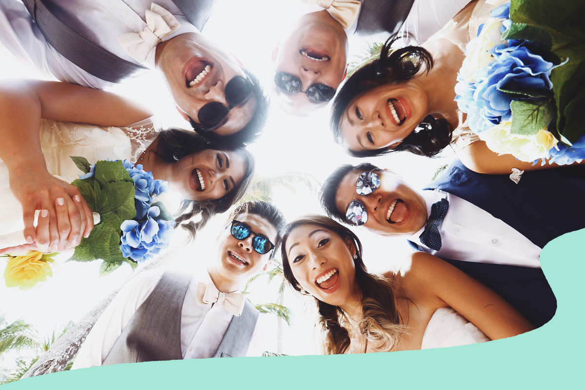 Wedding party gathered in a circle, smiling and looking down at the camera outdoors, with bouquets of blue and white flowers and palm trees visible in the background.