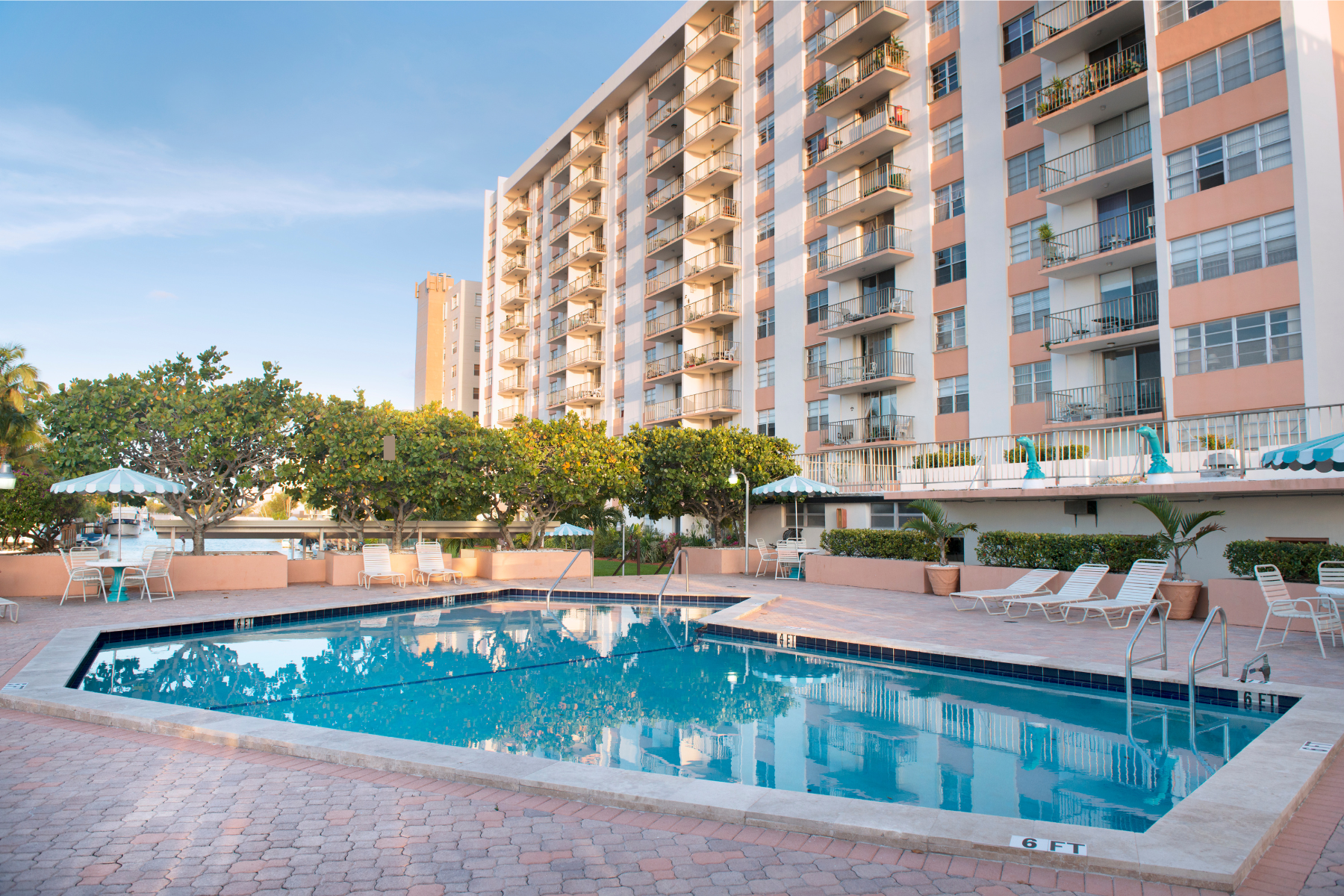 Residential condominium complex featuring a swimming pool, lounge chairs, and tropical landscaping under a clear blue sky.