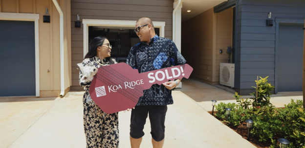 Two people stand in front of a newly built home holding a large ceremonial key that reads “Koa Ridge SOLD!” The pair pose in the driveway outside the residence, highlighting a home purchase or move-in celebration.