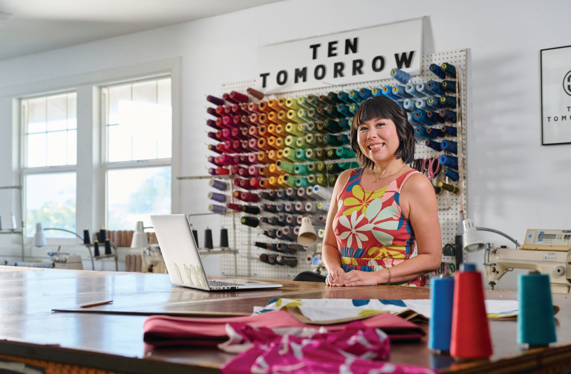Small business owner smiling in a sewing studio, surrounded by colorful thread, fabric, and sewing equipment, representing local entrepreneurship and craftsmanship.