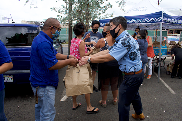 A community volunteer distributes a brown paper bag at an outdoor food distribution event, where several masked participants gather near tables and tents. Vehicles, volunteers, and supplies are visible throughout the busy parking lot setting.