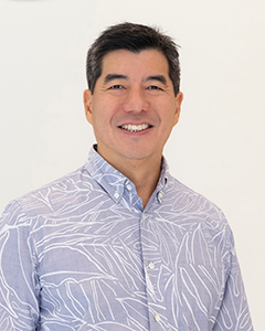 A professional headshot of a person wearing a light purple patterned aloha shirt, photographed against a plain white background.