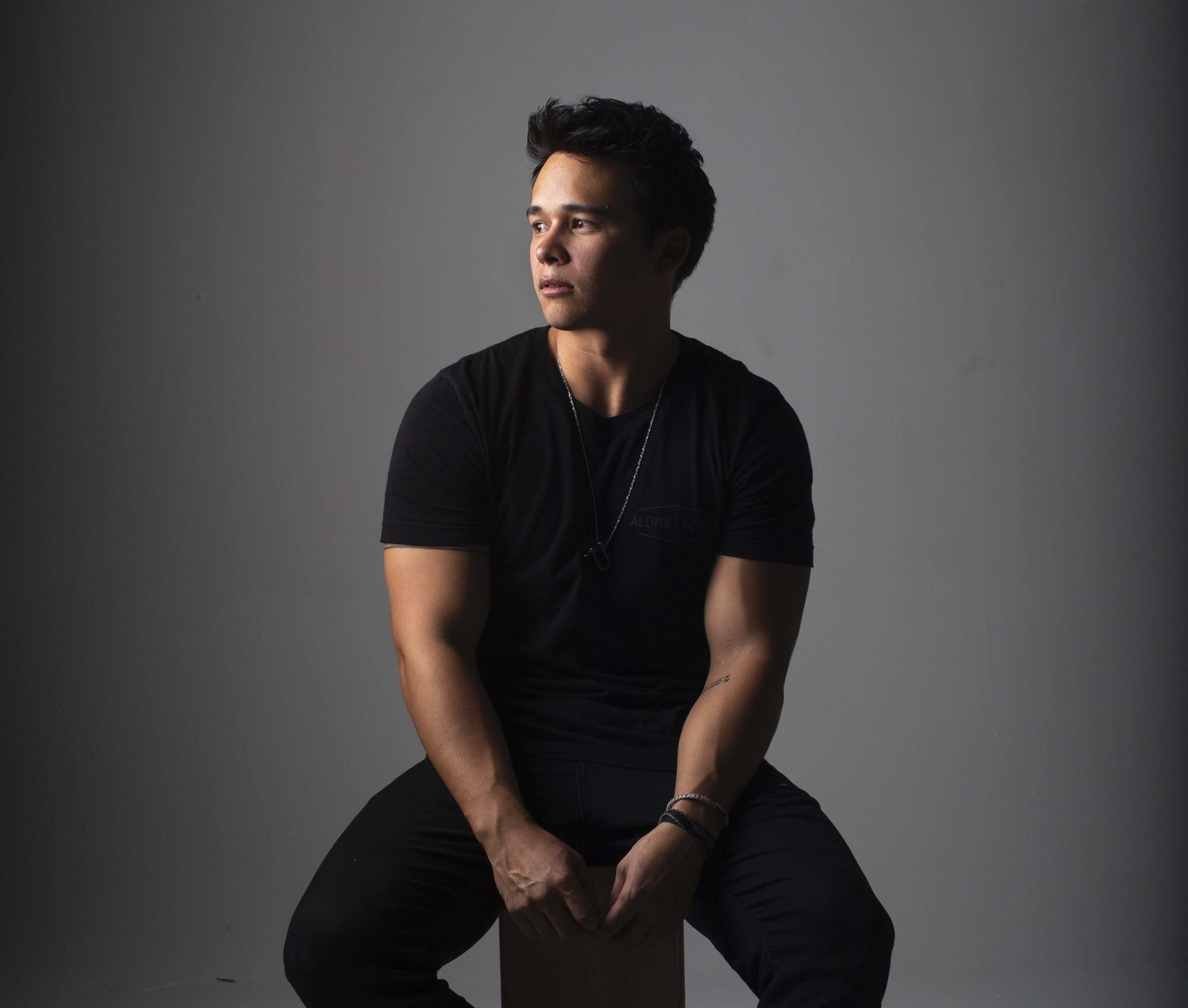 A studio portrait shows a person seated against a gray background, wearing a black T-shirt, dark pants, and a necklace. Dramatic side lighting highlights the subject while creating strong contrast and shadows.