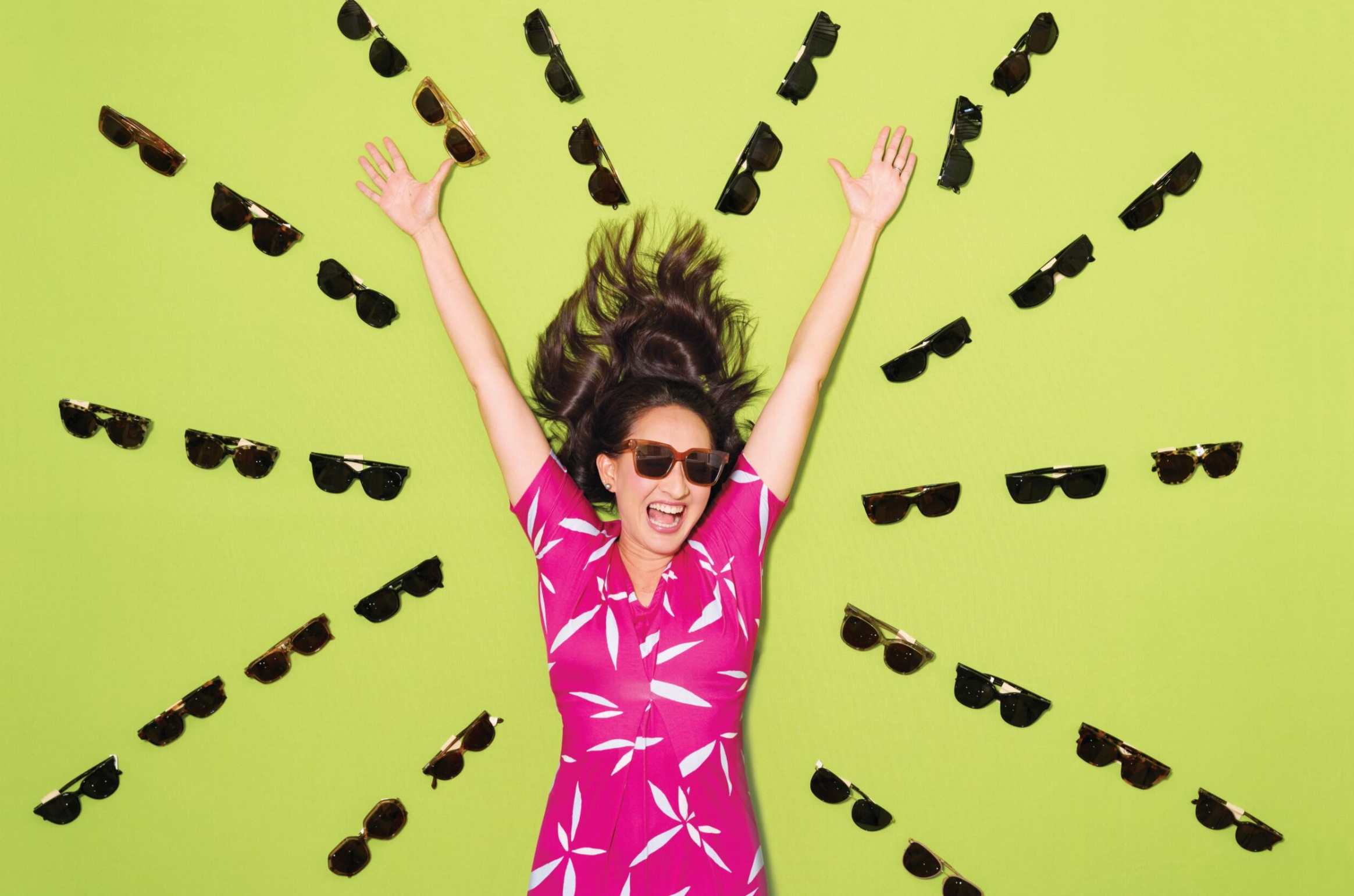 Person wearing sunglasses and a pink patterned outfit smiles with both arms raised while surrounded by an assortment of sunglasses arranged in a circular pattern on a bright green background.