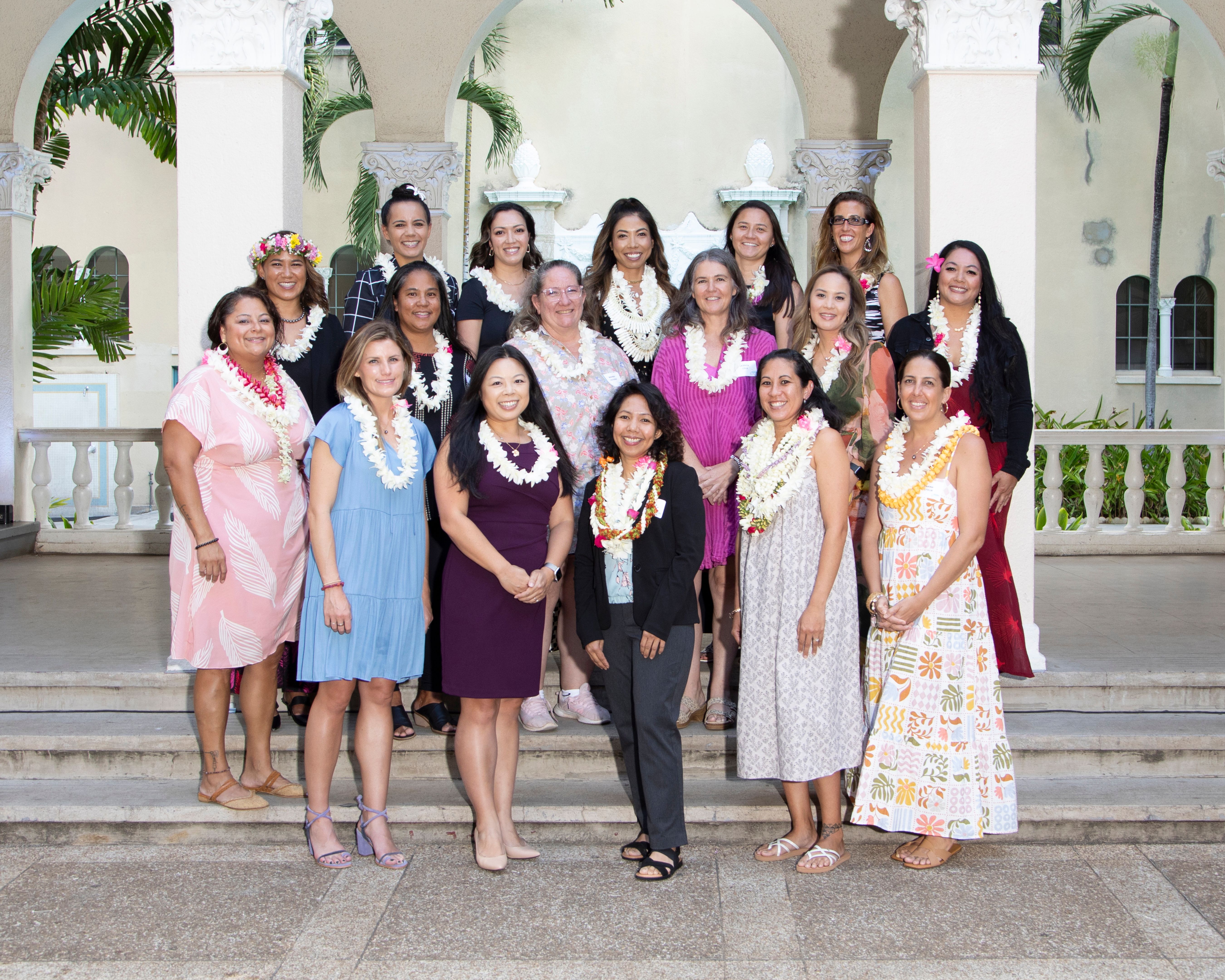 A group of people wearing flower leis pose for a group photo on the steps of an open-air courtyard with arches, columns, and tropical landscaping in the background. The gathering appears to be a professional or community event in a historic Hawaiian-style venue.