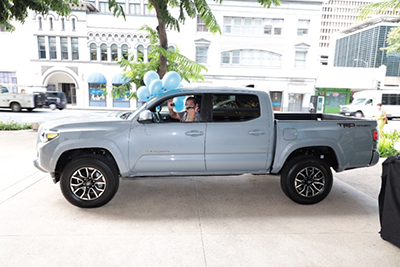 A light blue pickup truck decorated with blue balloons is parked in front of downtown buildings, with a person visible in the driver’s seat waving through the open window. The vehicle is displayed curbside beneath a tree-lined sidewalk during a promotional or community event.