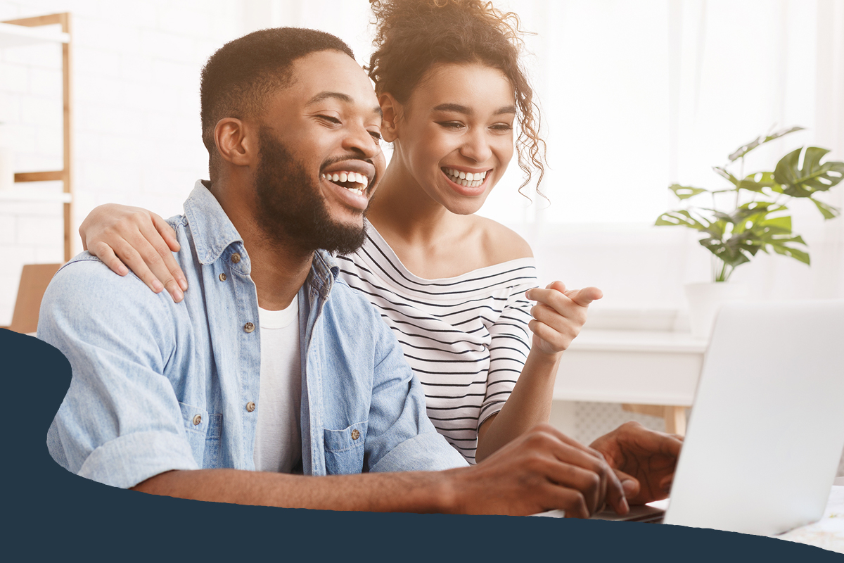 Two people smiling while using a laptop together at home.
