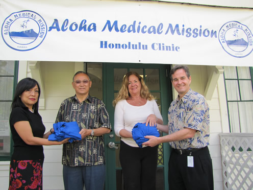 Four individuals pose in front of the Aloha Medical Mission Honolulu Clinic entrance while presenting donated blue supply bags.