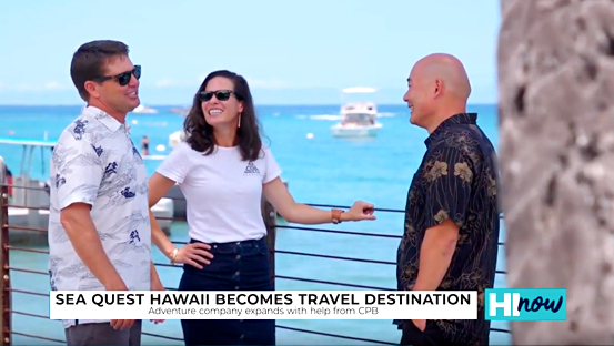 A still from a business feature segment showing three people talking on a waterfront promenade, with the ocean and boats in the background. On-screen text references Sea Quest Hawaii's expansion as a travel destination with support from Central Pacific Bank.