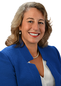 A professional headshot of a person wearing a royal blue blazer over a white blouse with a pearl necklace, photographed against a light gray background.