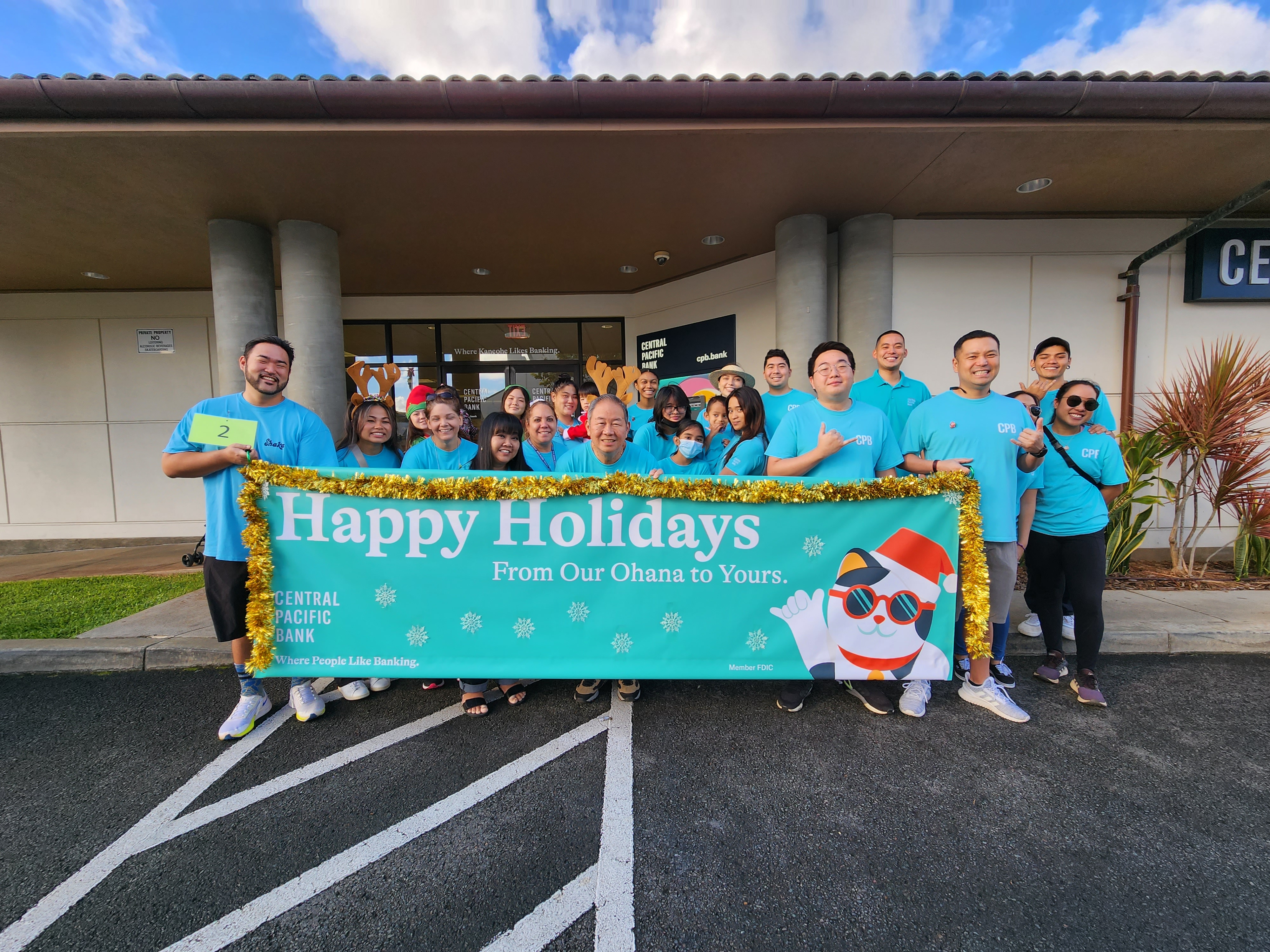 A group of Central Pacific Bank employees wearing matching teal shirts pose outside a bank branch holding a large banner that reads, “Happy Holidays From Our Ohana to Yours.” Several participants wear festive holiday accessories as they gather for a community holiday event.