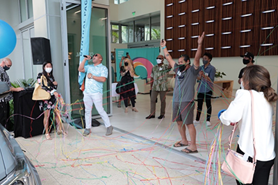 A group of people gathers in the Central Pacific Bank lobby during a celebration event, releasing colorful streamers while standing beneath a decorative wood wall installation. Several attendees wear face masks as they participate in the festive ribbon-cutting or unveiling ceremony.  Provide your feedback on BizChat