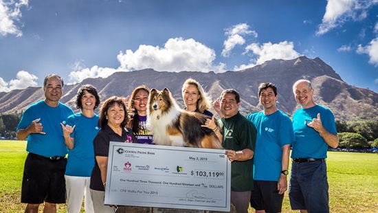 A group of people stand outdoors with a Collie dog and a large Central Pacific Bank donation check, posing in a grassy field with mountains and a blue sky in the background.